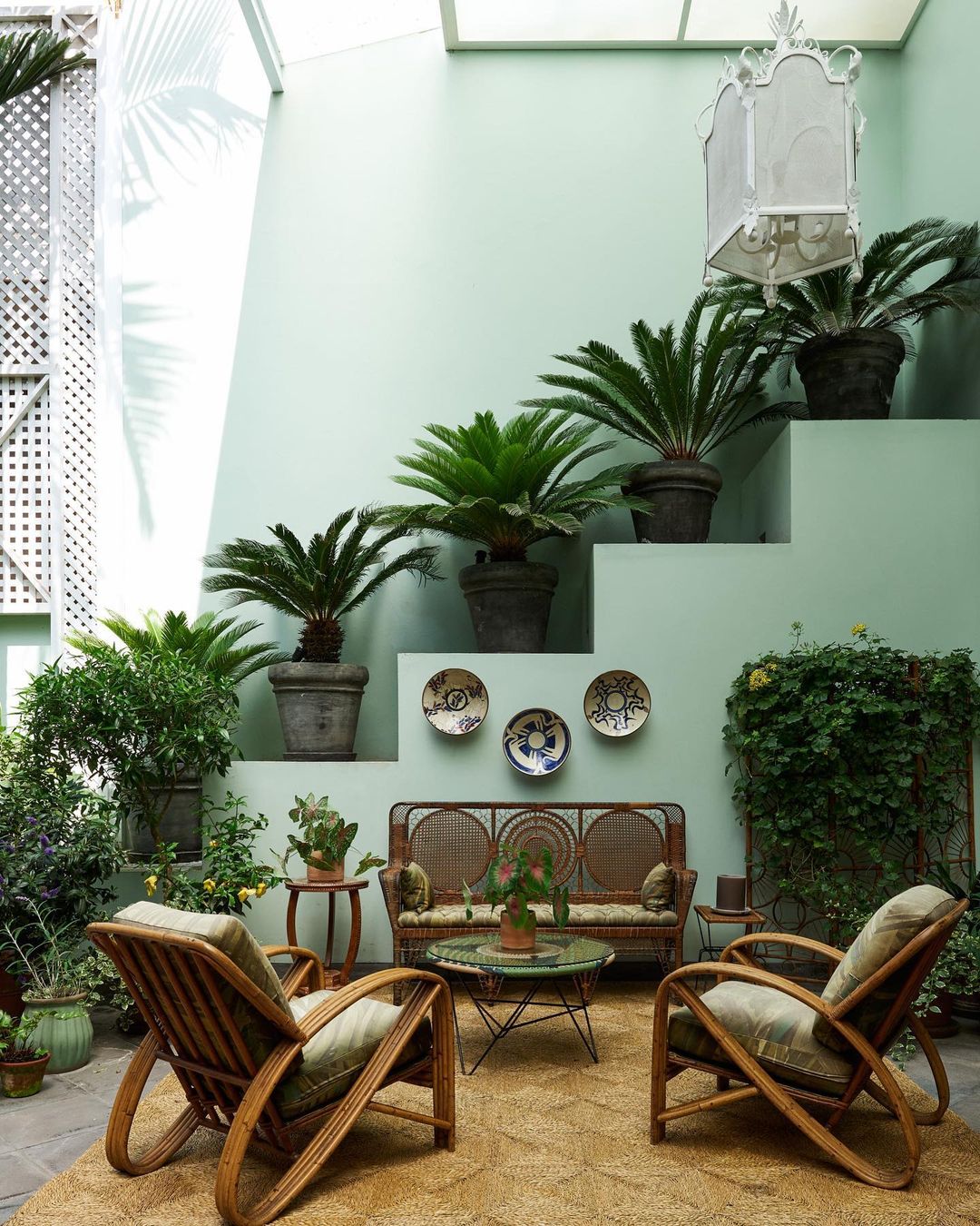 Rattan seating arrangement with a sofa and two armchairs on a jute rug, surrounded by potted plants, beneath a skylight, indoors.