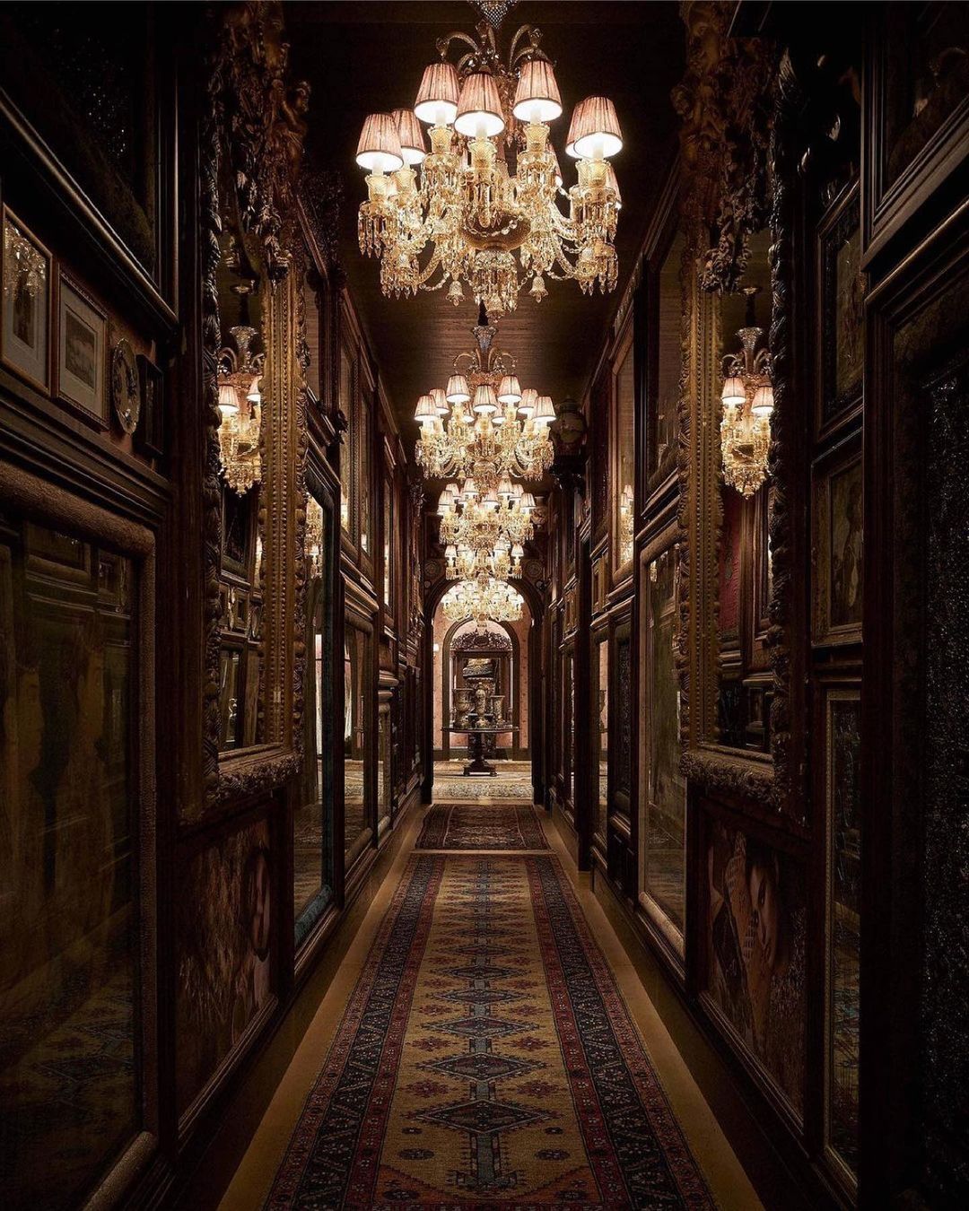 Opulent hallway featuring multiple crystal chandeliers, framed art, mirrors, and a patterned carpet in an elegant, vintage setting.