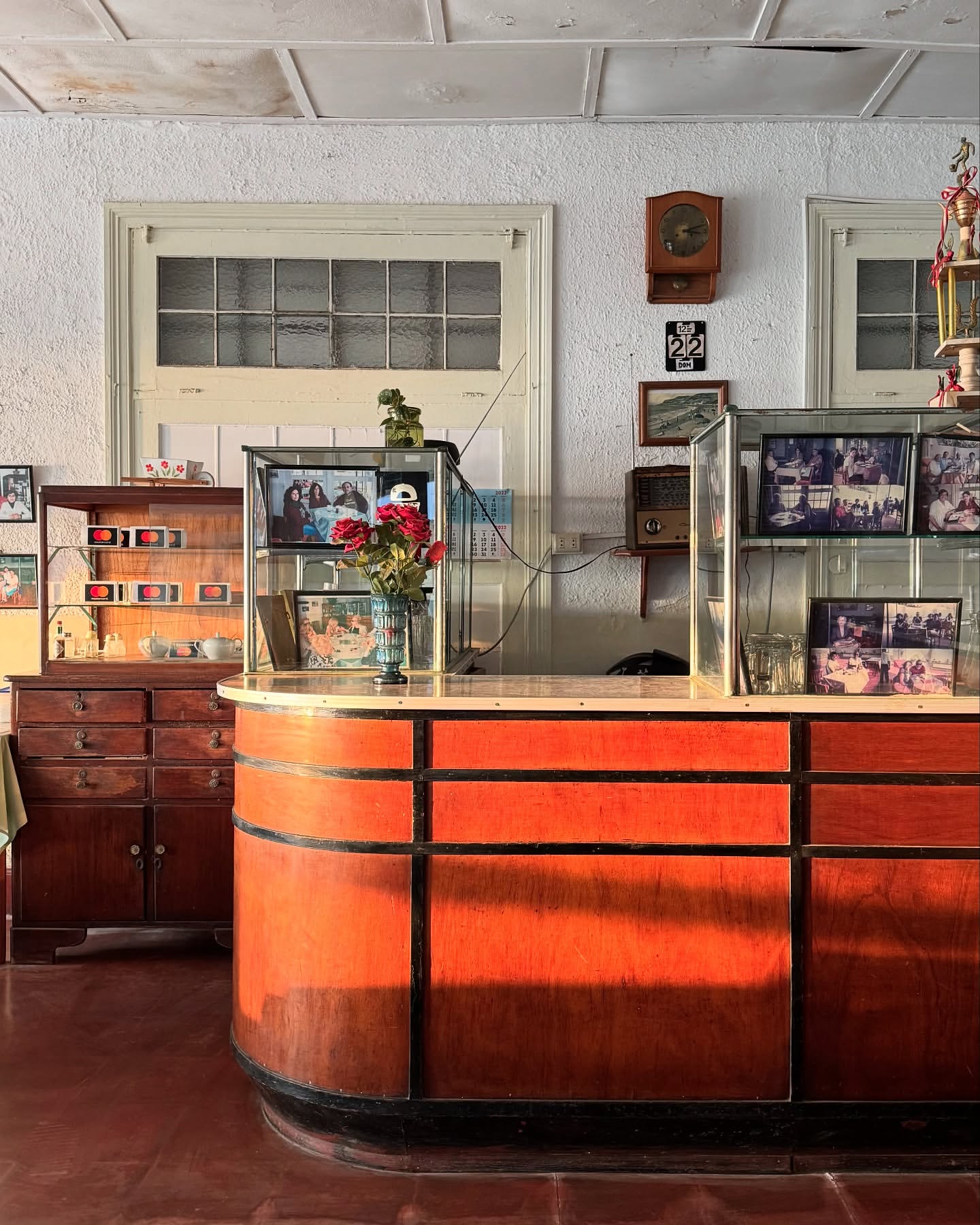 Vintage hotel lobby with orange reception desk, glass displays, clock, pictures, and tiled floor in a warm, inviting setting.