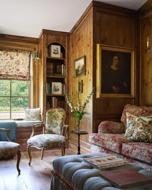 Warm, wood-paneled room features a floral sofa, upholstered armchair, ottoman, and a window seat, complemented by books and artwork.