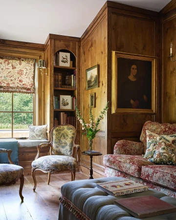 Warm, wood-paneled room features a floral sofa, upholstered armchair, ottoman, and a window seat, complemented by books and artwork.