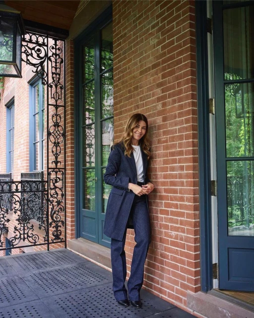 A woman in a dark blue suit leans against a brick wall outside, with decorative ironwork and greenery visible in the background.