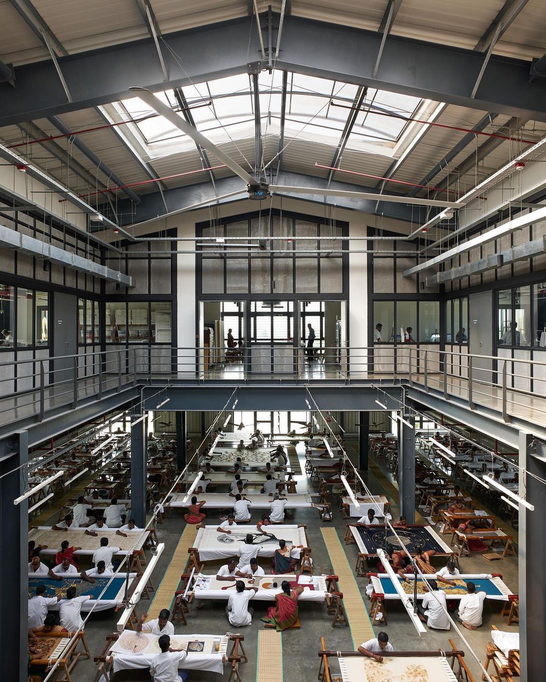 High-angle shot inside a large workshop, showing people seated at tables working on textiles, with an upper-level overlooking the scene.