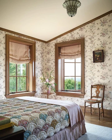 Traditional bedroom with floral wallpaper, wooden trim, two windows, a quilted bed, and a wooden chair photographed by Bjorn Wallander.