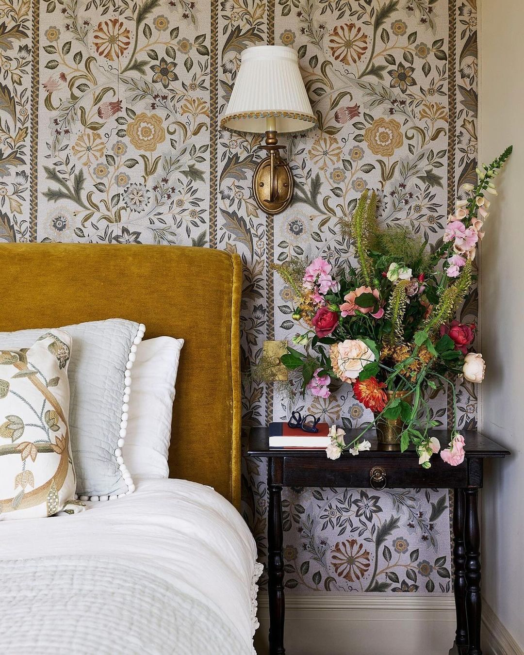 Bedroom interior featuring a bed with a mustard yellow headboard, floral wallpaper, and a bouquet of flowers on a bedside table.