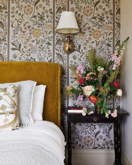 Bedroom interior featuring a bed with a mustard yellow headboard, floral wallpaper, and a bouquet of flowers on a bedside table.