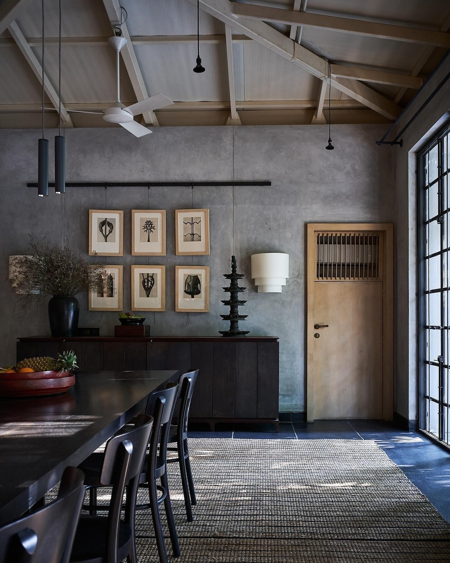 Dining room interior featuring a dark wood table and chairs, a grey textured wall with framed prints, and natural light from windows.
