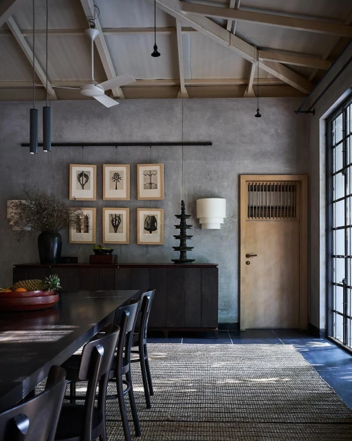 Dining room interior featuring a dark wood table and chairs, a grey textured wall with framed prints, and natural light from windows.