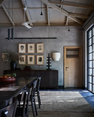 Dining room interior featuring a dark wood table and chairs, a grey textured wall with framed prints, and natural light from windows.