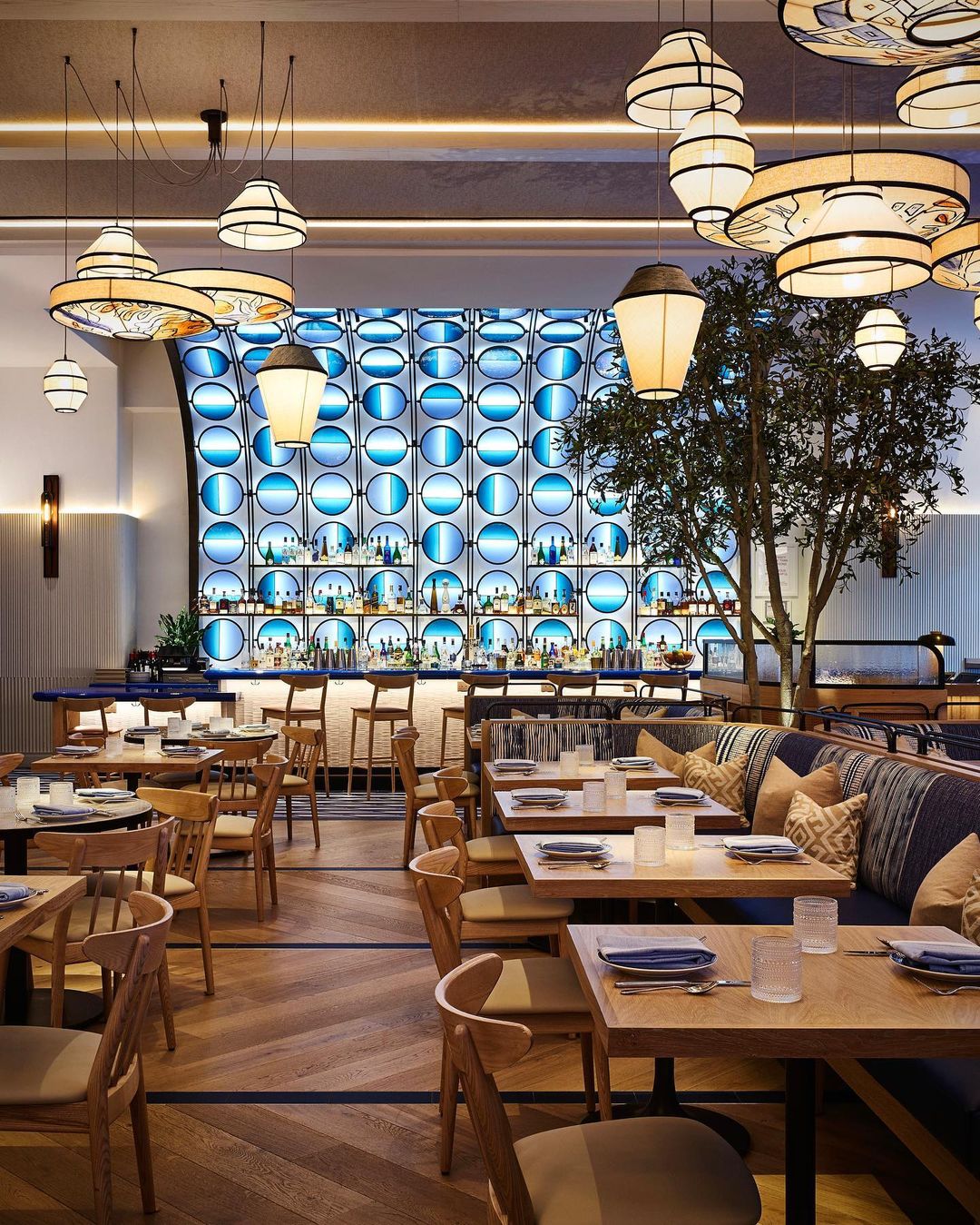 Upscale restaurant interior featuring wooden tables, chairs, lantern lighting, and a bar with blue decorative backdrop.