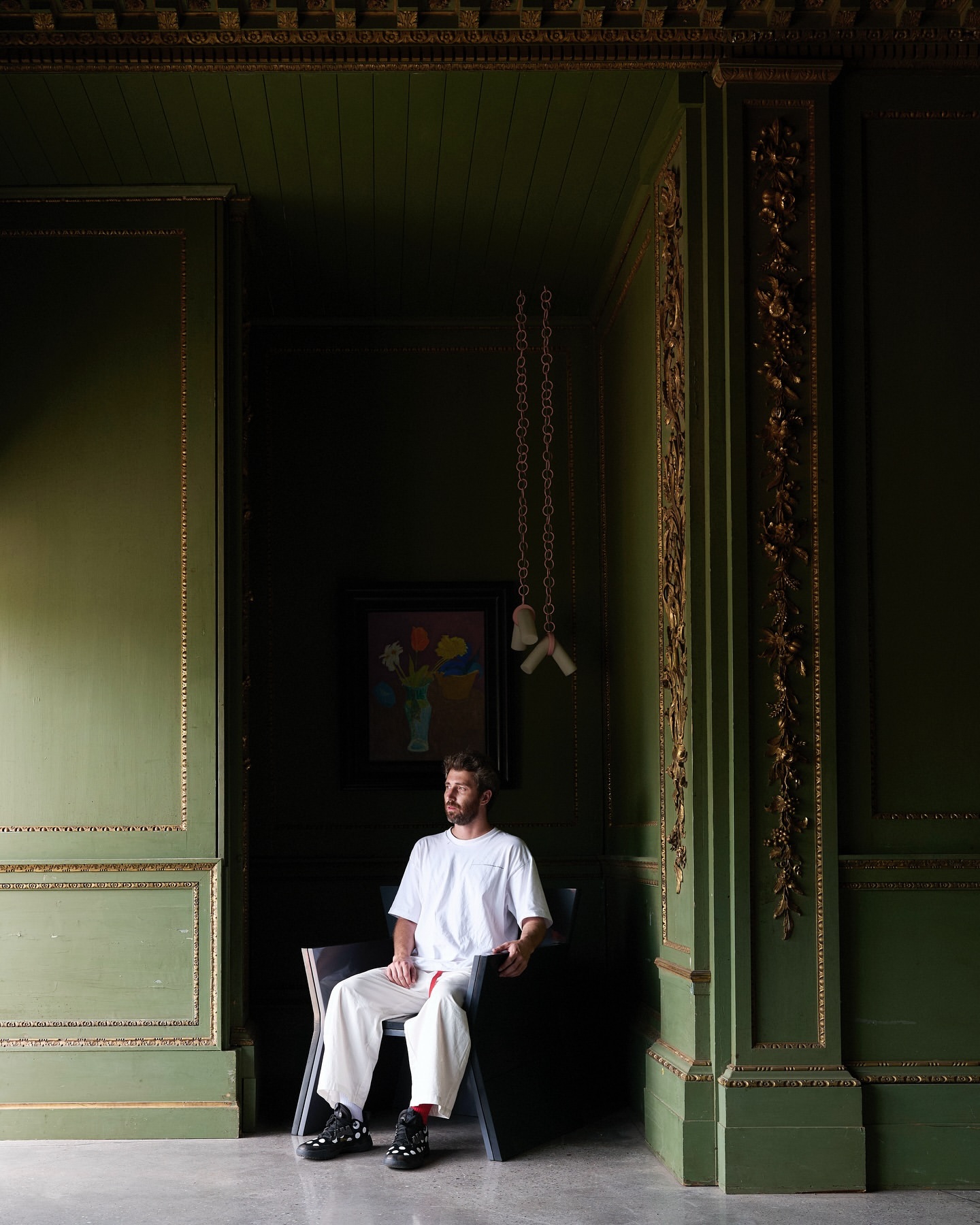 Bearded man in white t-shirt and white trousers seated on dark chair in dark green room with gilded architectural wall paneling