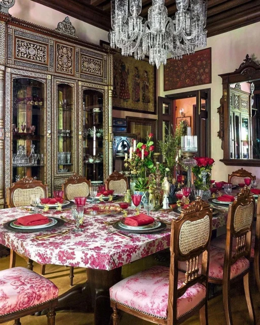 Ornate dining room featuring a long table set with china and red napkins, surrounded by upholstered chairs, lit by a crystal chandelier.