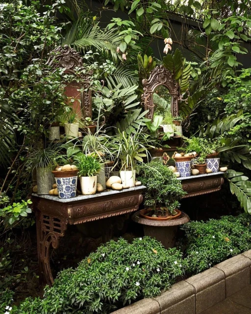 Indoor garden display featuring ornate metal tables holding potted plants and stone pebbles surrounded by lush green foliage.