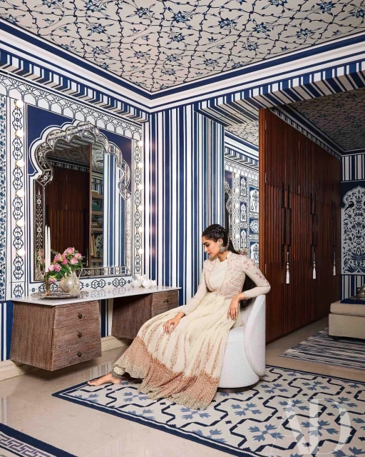 An adult woman sits in a designer chair indoors in a room with blue and white wallpaper, a table with flowers, and hardwood floors.
