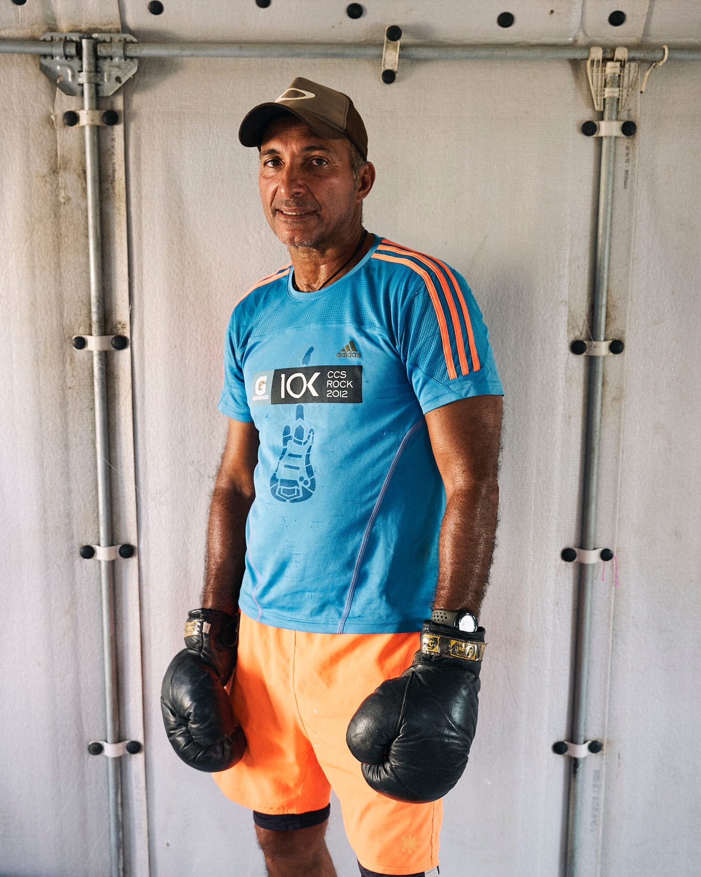 Adult male posing in a photo studio wearing boxing gloves and athletic wear, a casual hat, and standing against a white wall.