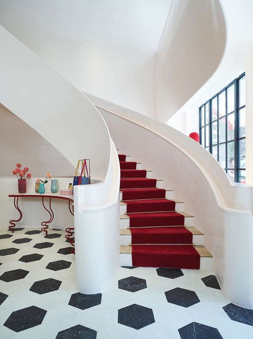 Interior view of a curved staircase with a red carpet, a table with floral arrangements, and a geometric tiled floor.