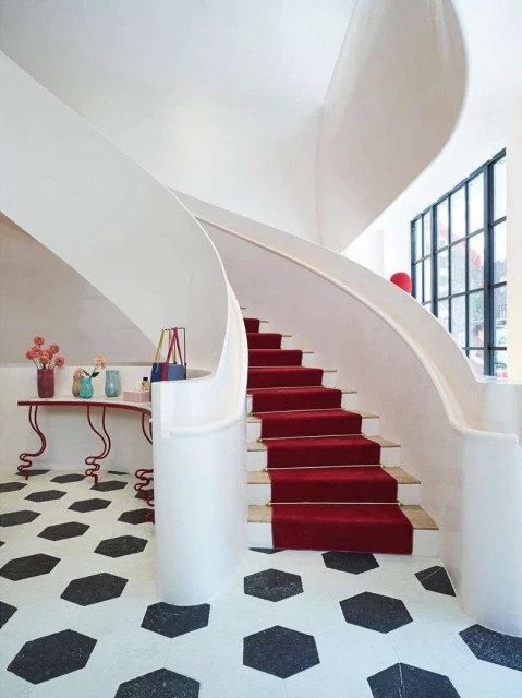 Interior view of a curved staircase with a red carpet, a table with floral arrangements, and a geometric tiled floor.