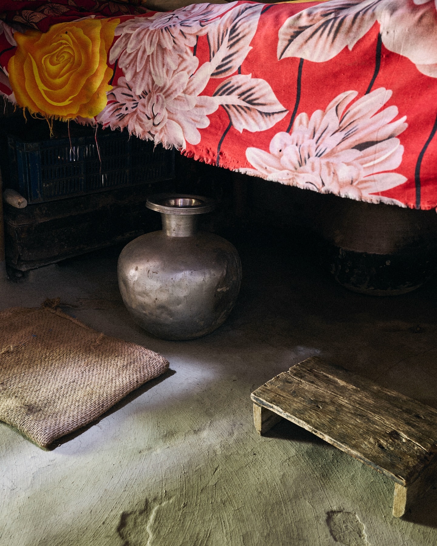 Still life featuring a metal water pot, a woven mat, and a wooden footstool on a textured cement floor with a floral bedspread backdrop.