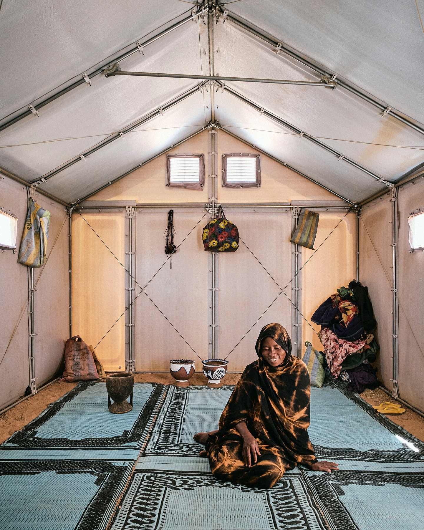 Woman sits inside a white canvas tent on a patterned mat. Bags hang on the walls of the tent, which is lit by natural light.