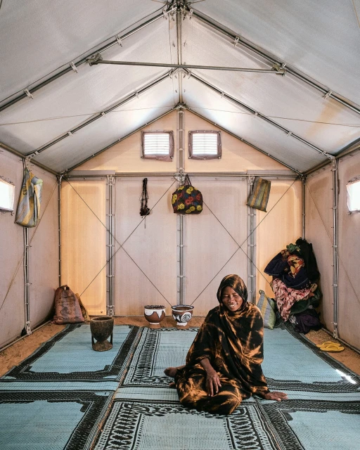 Woman sits inside a white canvas tent on a patterned mat. Bags hang on the walls of the tent, which is lit by natural light.