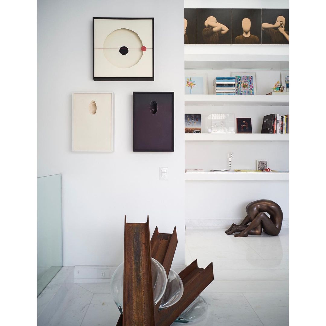 Interior view of a room with abstract art on the walls, a clear glass railing, modern sculpture, white floors and walls, and natural lighting.