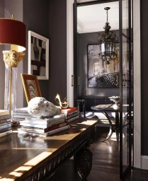 Dark interior scene with a wooden console table holding stacked books, a white stone bust, and an ornate table lamp with a red shade.