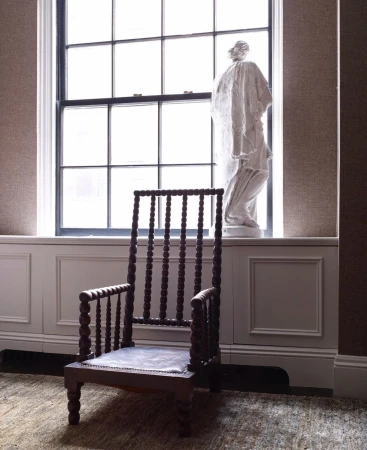 A wooden spindle chair sits on a textured rug in a room with a window, white radiator cover, and a classical bust sculpture on the sill.