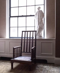 A wooden spindle chair sits on a textured rug in a room with a window, white radiator cover, and a classical bust sculpture on the sill.