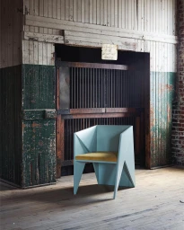 Modern geometric light blue armchair with a yellow cushion positioned on a weathered wooden floor inside an industrial factory.