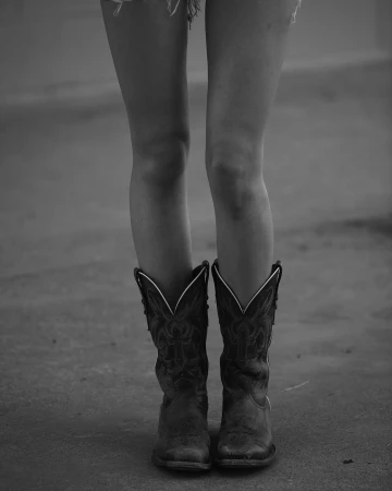 Close-up of a person's legs wearing embroidered dark leather cowboy boots and frayed denim shorts.