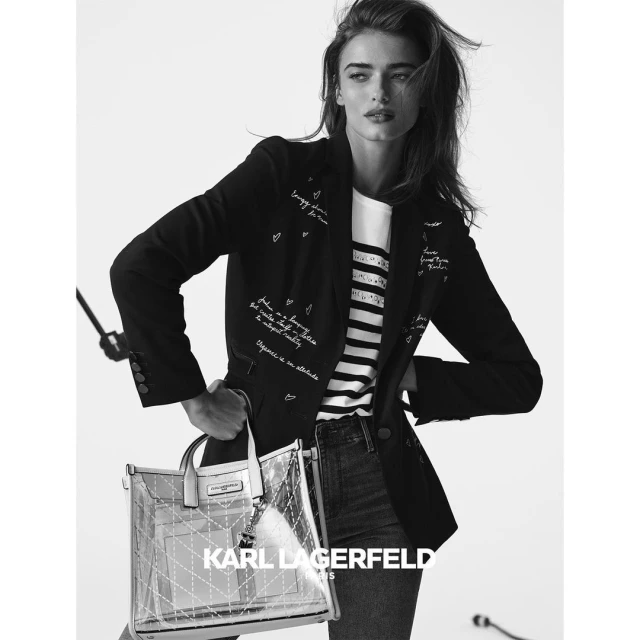Black-and-white fashion shot of a woman in a blazer holding a tote bag. She is posing in a light studio setting with a bright white background.