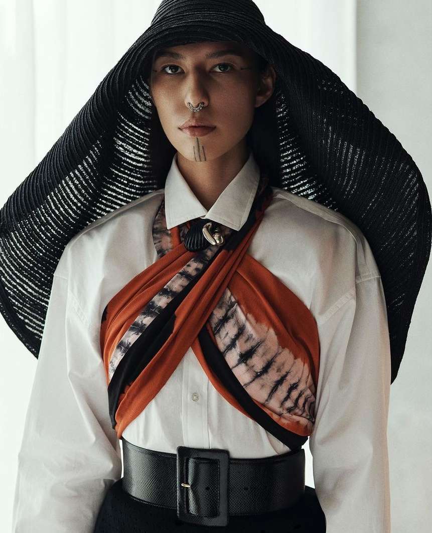 Model poses in a white button-down shirt, belted black skirt, rust-colored scarf, and a wide-brimmed black hat in a studio setting.