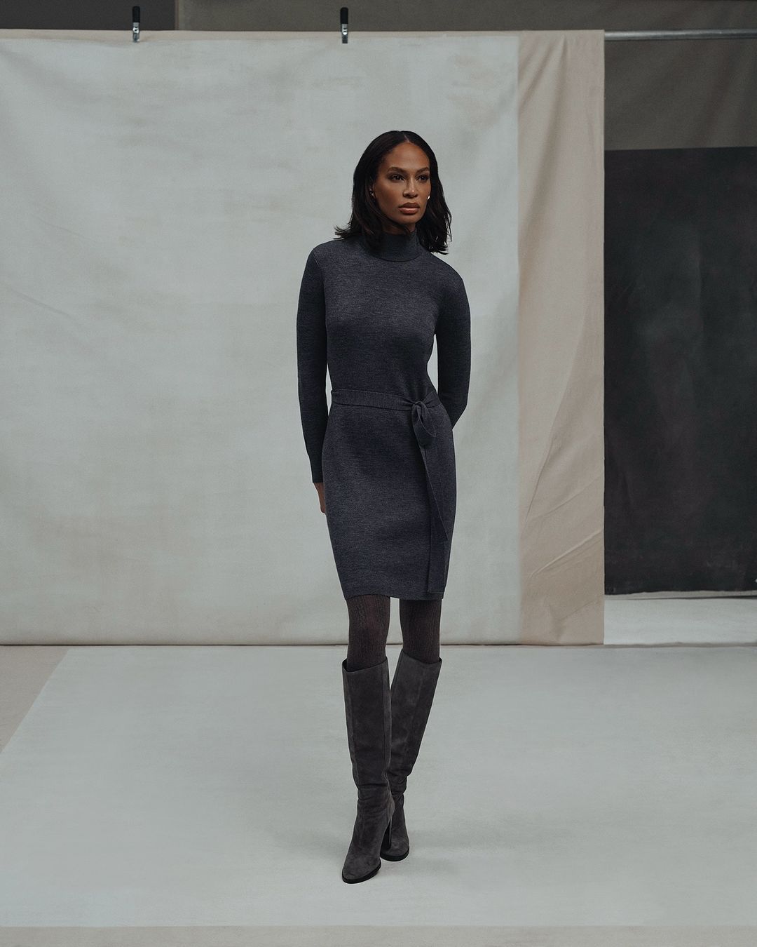 A woman in a gray turtleneck dress and knee-high boots poses against a neutral backdrop in a studio setting.