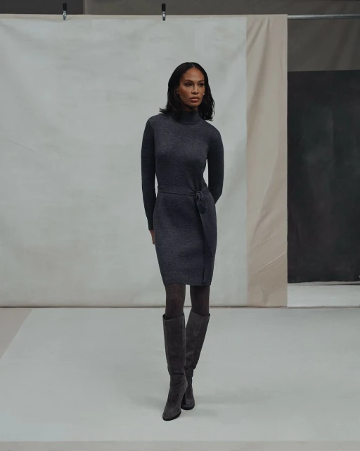 A woman in a gray turtleneck dress and knee-high boots poses against a neutral backdrop in a studio setting.
