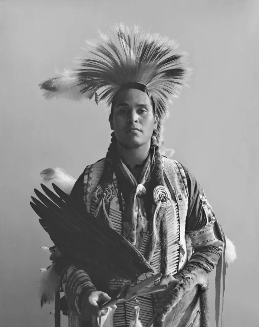Portrait of indigenous man in traditional headdress and clothing holding an eagle wing against a neutral backdrop in black and white.