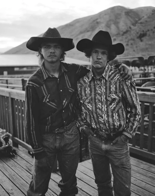 Black and white portrait of two teenage cowboys standing shoulder to shoulder on a wooden platform with a mountain in the background.