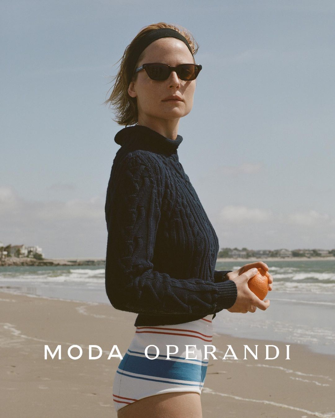A woman in a navy sweater holds an orange fruit on a beach with water and sky in the background, shot in natural light.