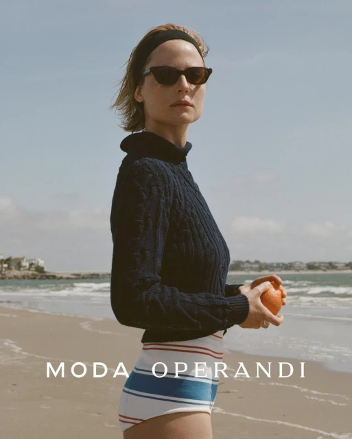 A woman in a navy sweater holds an orange fruit on a beach with water and sky in the background, shot in natural light.