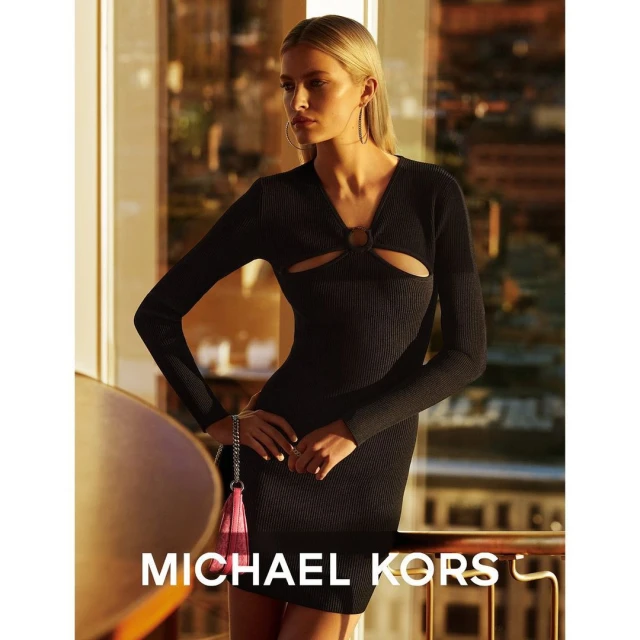 Fashion shot of blonde woman in a black ribbed long-sleeve dress with cutouts, holding a pink fringed purse in a city apartment.