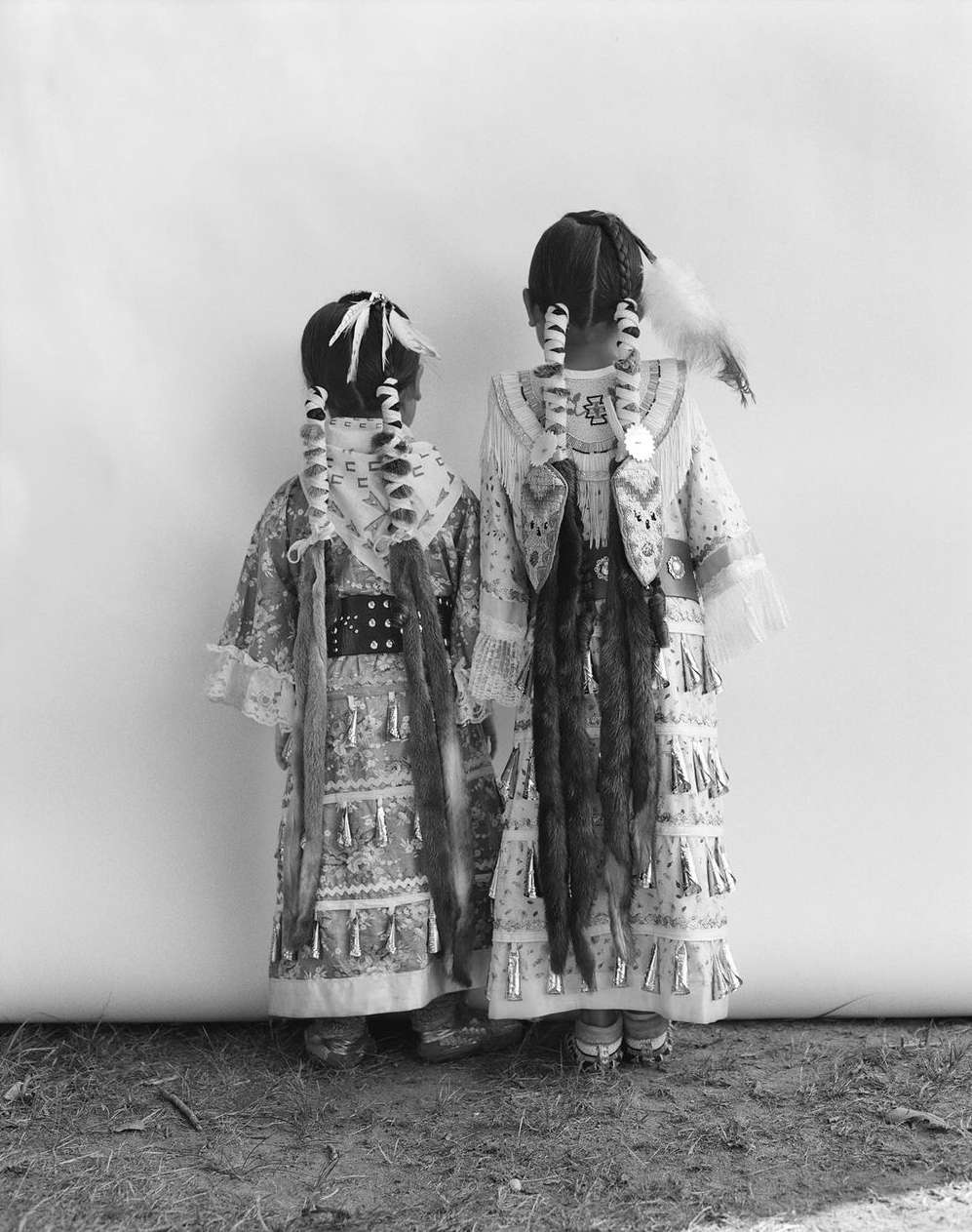 Two children in traditional Native American clothing stand back to back against a white wall in a black and white outdoor photograph.