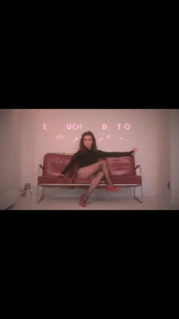 A woman in a black bodysuit and red high heels sits on a red couch in front of a white wall with neon lettering.
