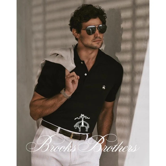 A man with curly hair and sunglasses poses in a black polo shirt and white pants, exuding a casual yet sophisticated style indoors.