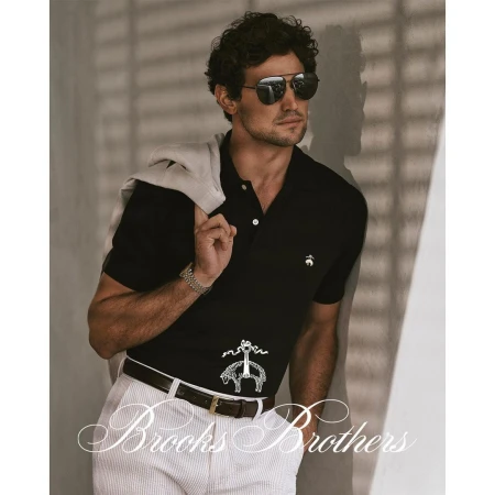 A man with curly hair and sunglasses poses in a black polo shirt and white pants, exuding a casual yet sophisticated style indoors.