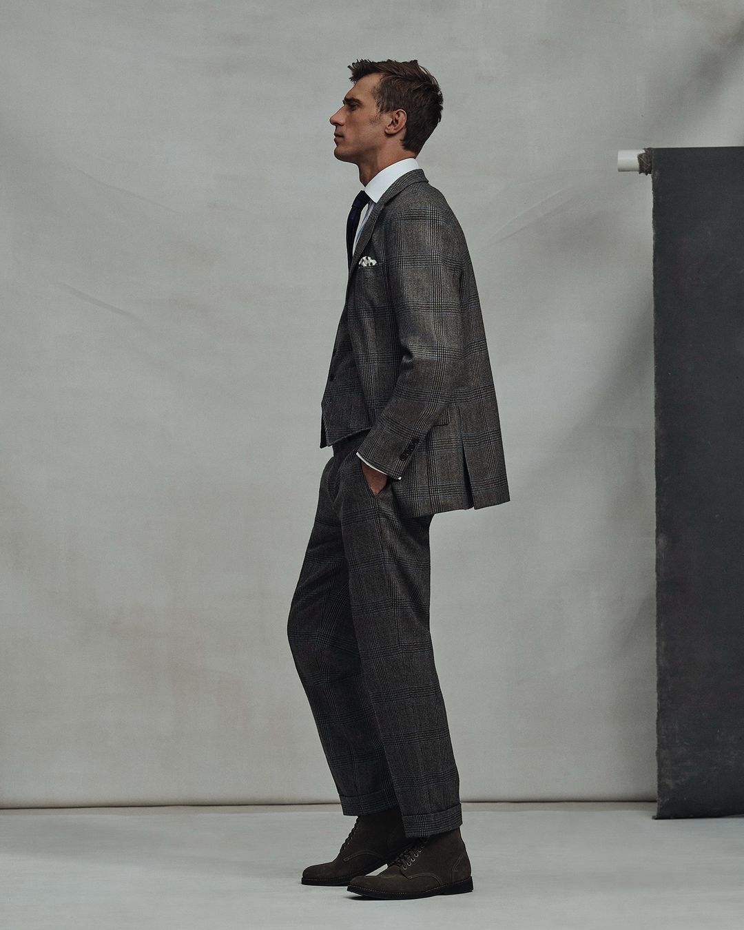 A man in a grey suit stands in profile in a studio, showcasing tailored menswear. The ensemble includes a jacket, trousers, and dress shoes.