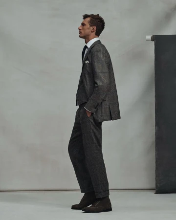 A man in a grey suit stands in profile in a studio, showcasing tailored menswear. The ensemble includes a jacket, trousers, and dress shoes.