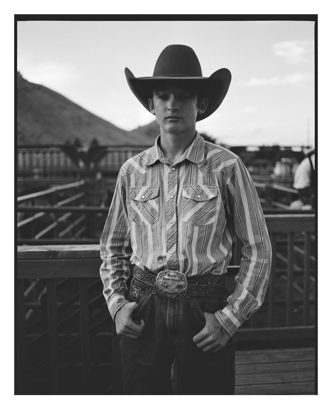 Black-and-white portrait of a teenage boy in a cowboy hat and western shirt, standing in front of a mountain landscape.