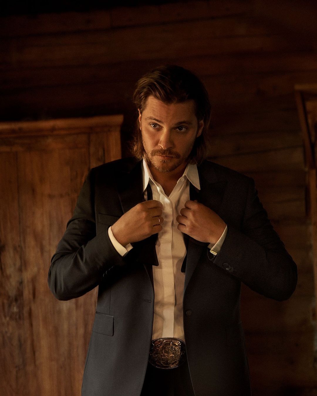 Man in black suit adjusts tie in rustic setting, showcasing modern Western fashion with a hint of old-world charm and rich wooden textures.