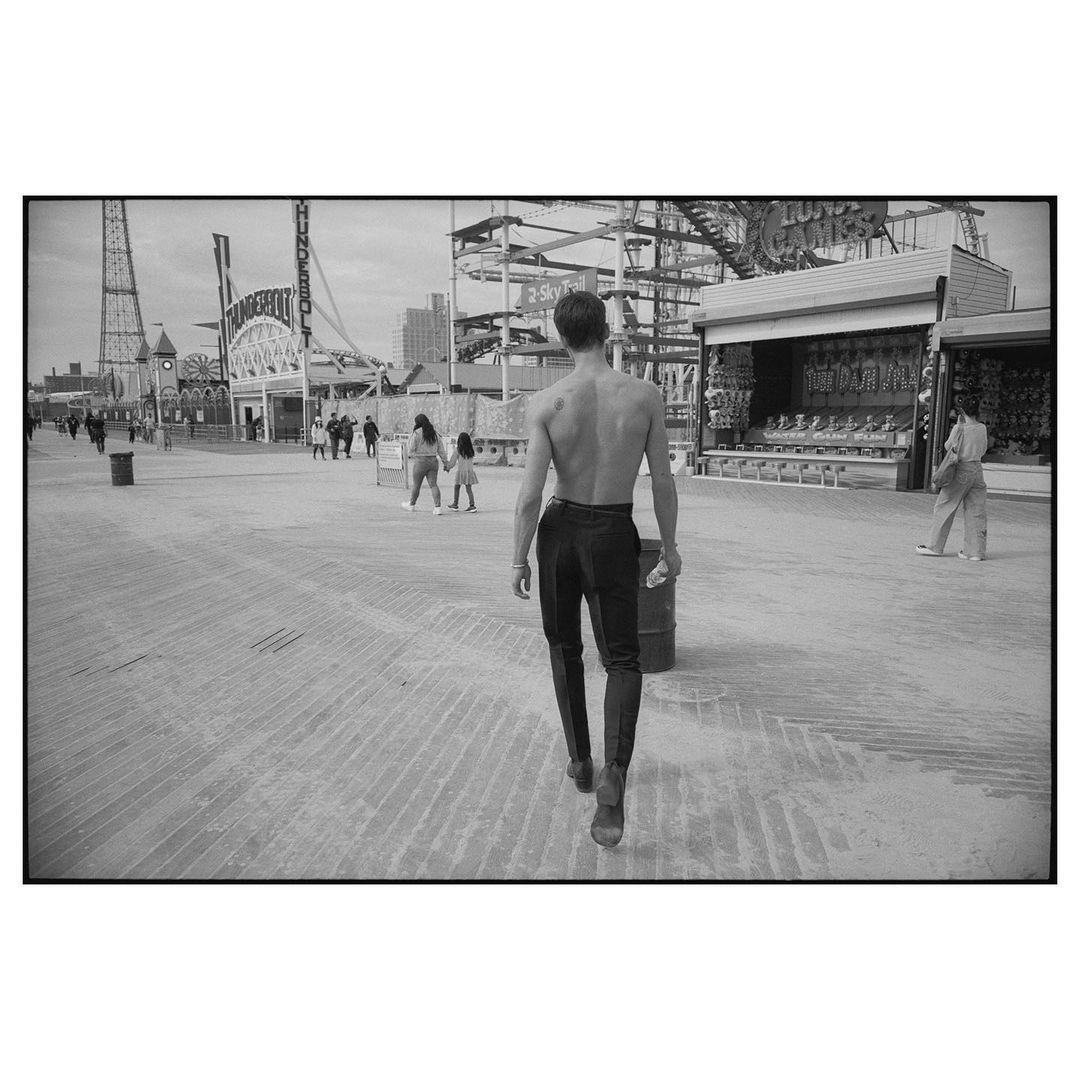 Black and white photograph of shirtless man walking toward amusement park boardwalk with game stalls and roller coaster in background.