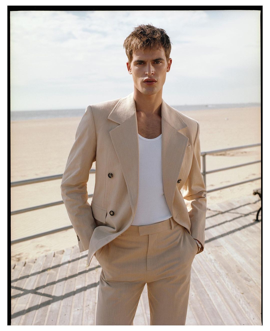 A blond male model in a beige suit poses outdoors on a sunny day, with the beach and horizon visible in the background.