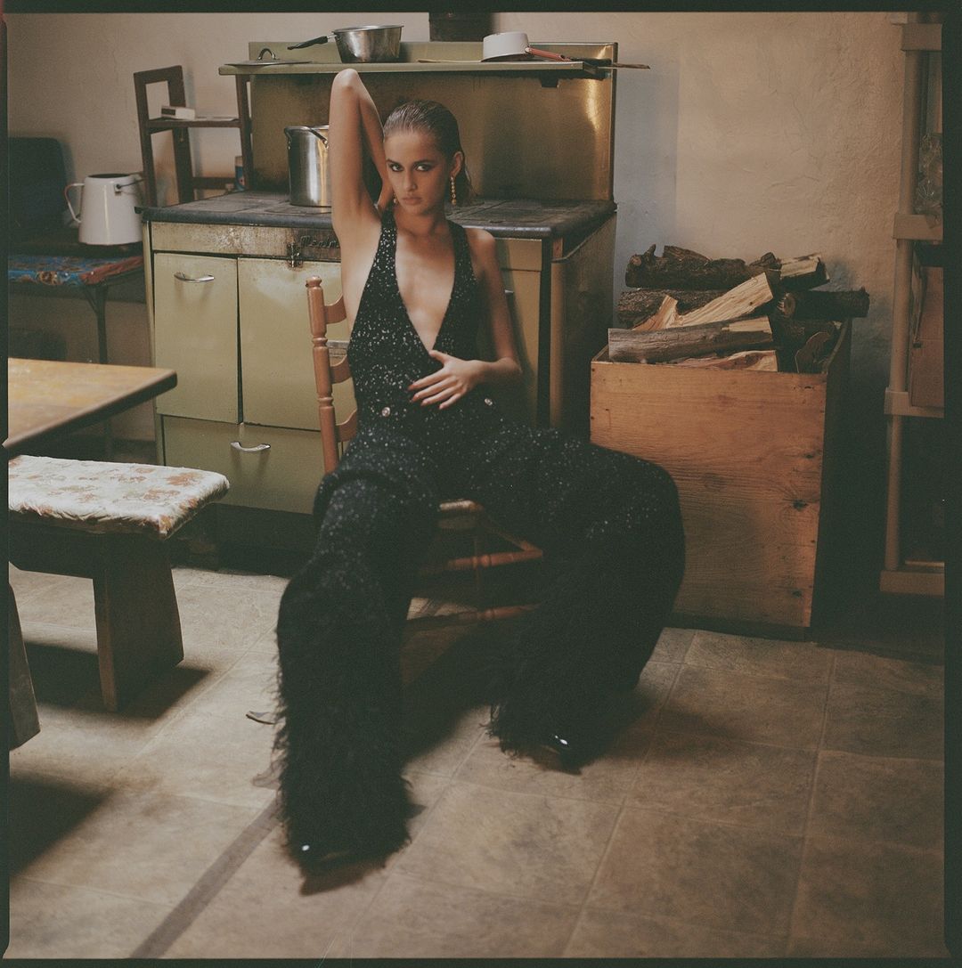 A stylish woman with dark hair sits in a chair wearing a shimmering black jumpsuit in a vintage-style room with rustic decor.